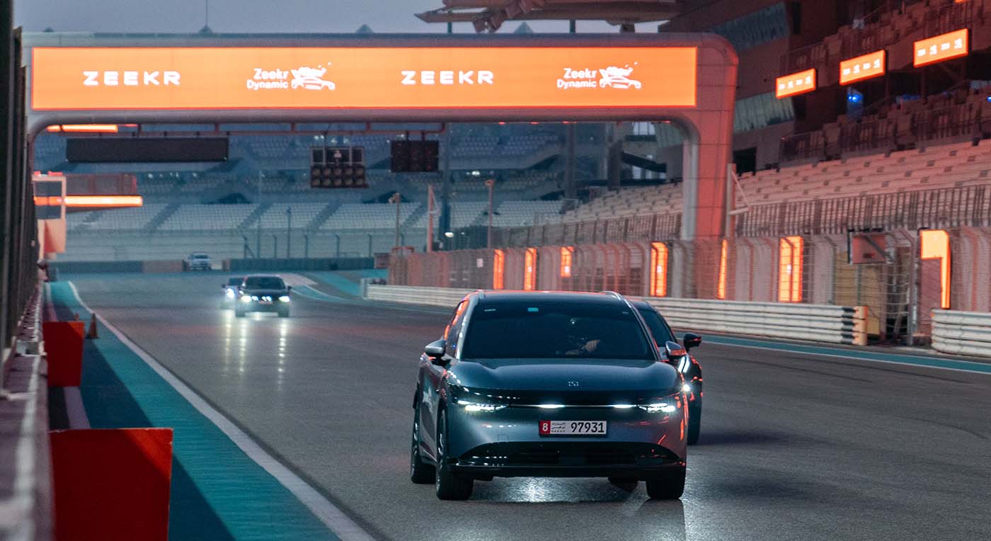 Sleek dark grey electric sedan leading a pack of performance cars down a race track below a orange Zeekr arch.